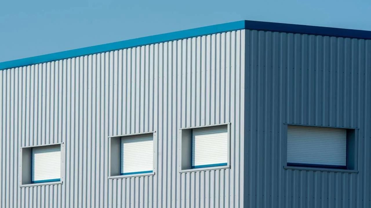 Corner of a corrugated-metal industrial warehouse with shuttered windows under a clear sky.
