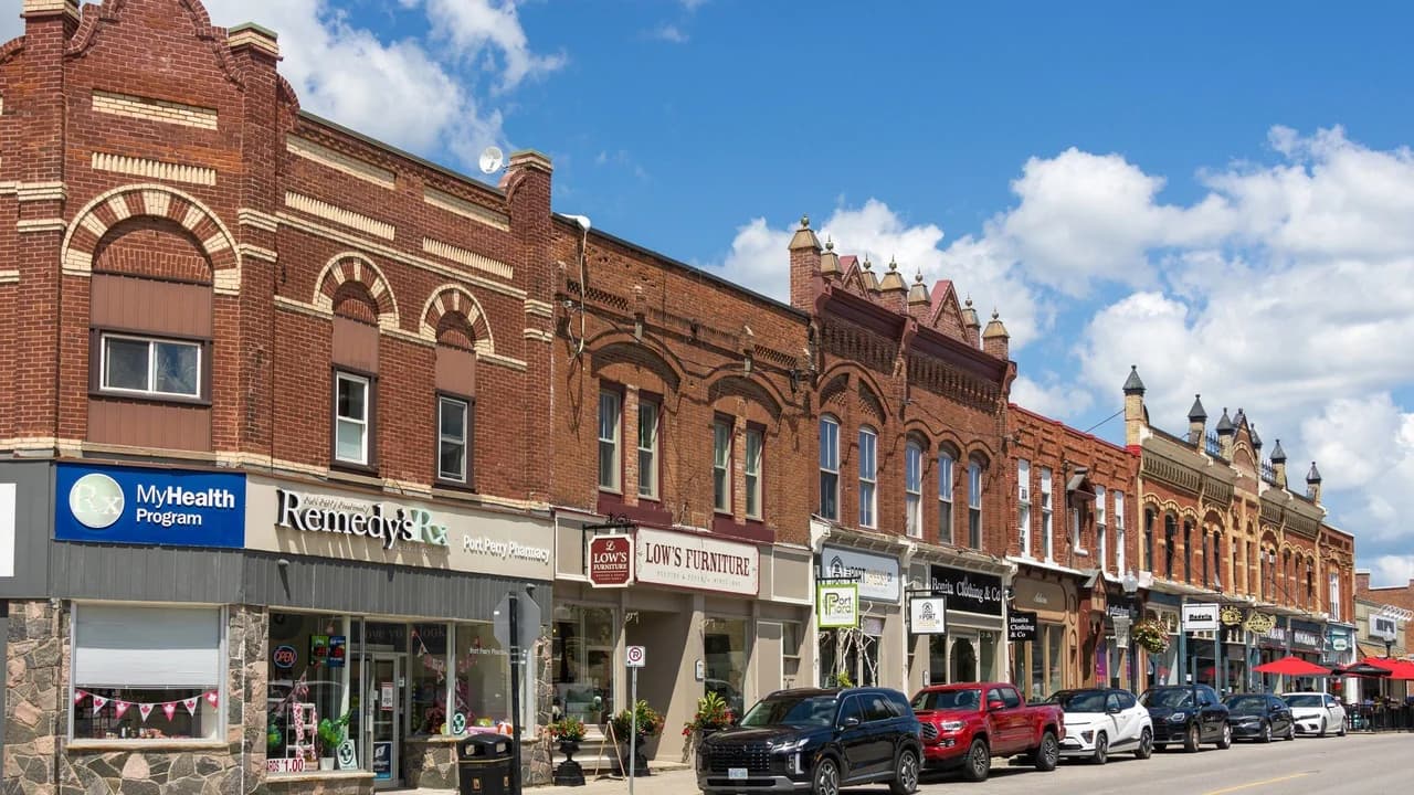 Historic brick storefronts on Queen Street in downtown Port Perry, Ontario, with parked cars under a blue sky.